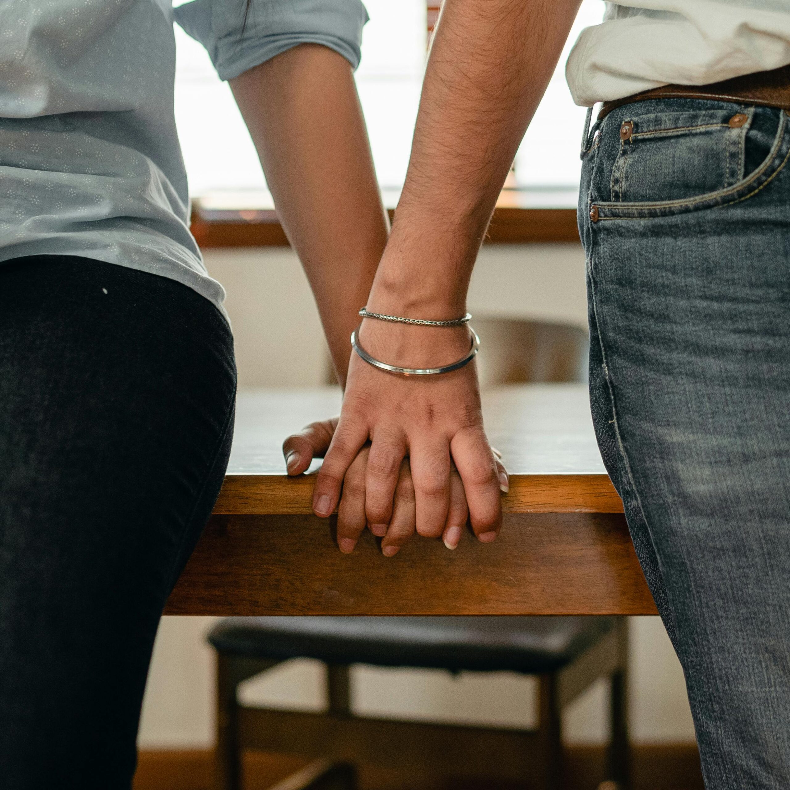 Crop anonymous young romantic couple in shirts and jeans holding hands while leaning on wooden table